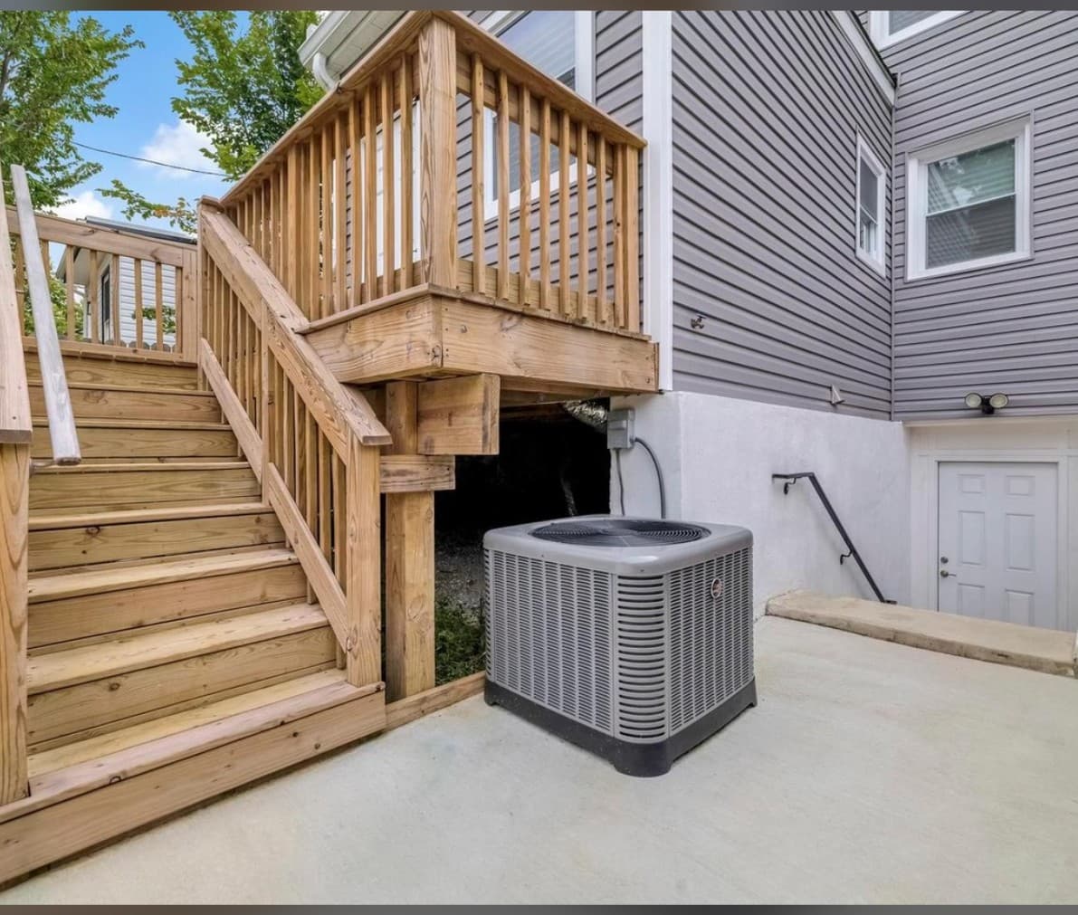 Outdoor air conditioning unit on a concrete pad beside wooden stairs and a grey house.